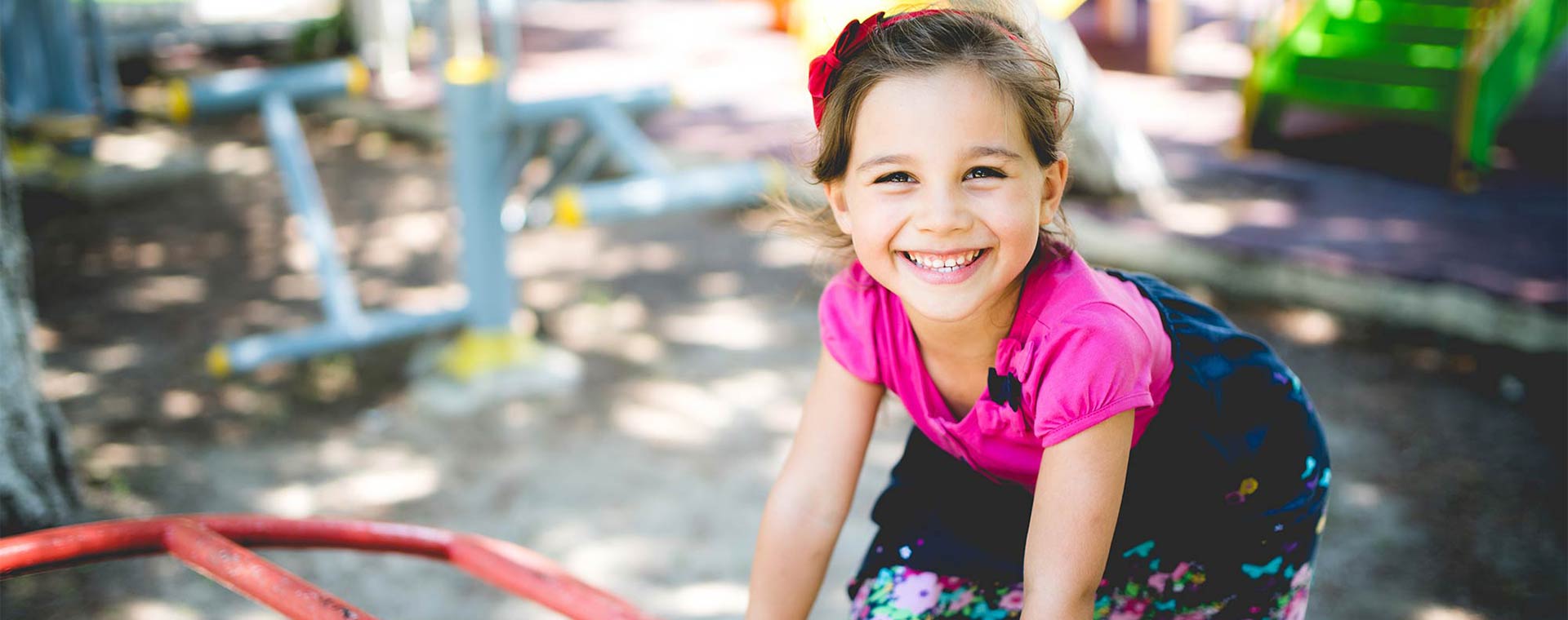 A smiling young girl is climbing on equipment at a playground. She is wearing a bright pink top and a navy blue skirt, with a red bow in her hair.