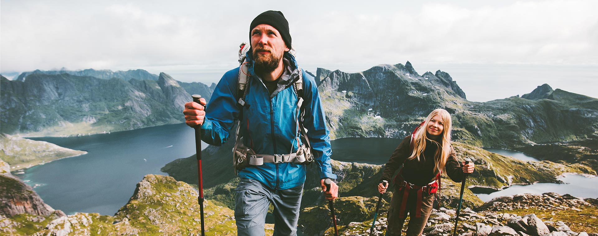 A man and a woman are hiking in the mountains, they have reached a summit and behind them is a lake. They are both wearing rain coats and large backpacks. They are using poles to assist in walk.
