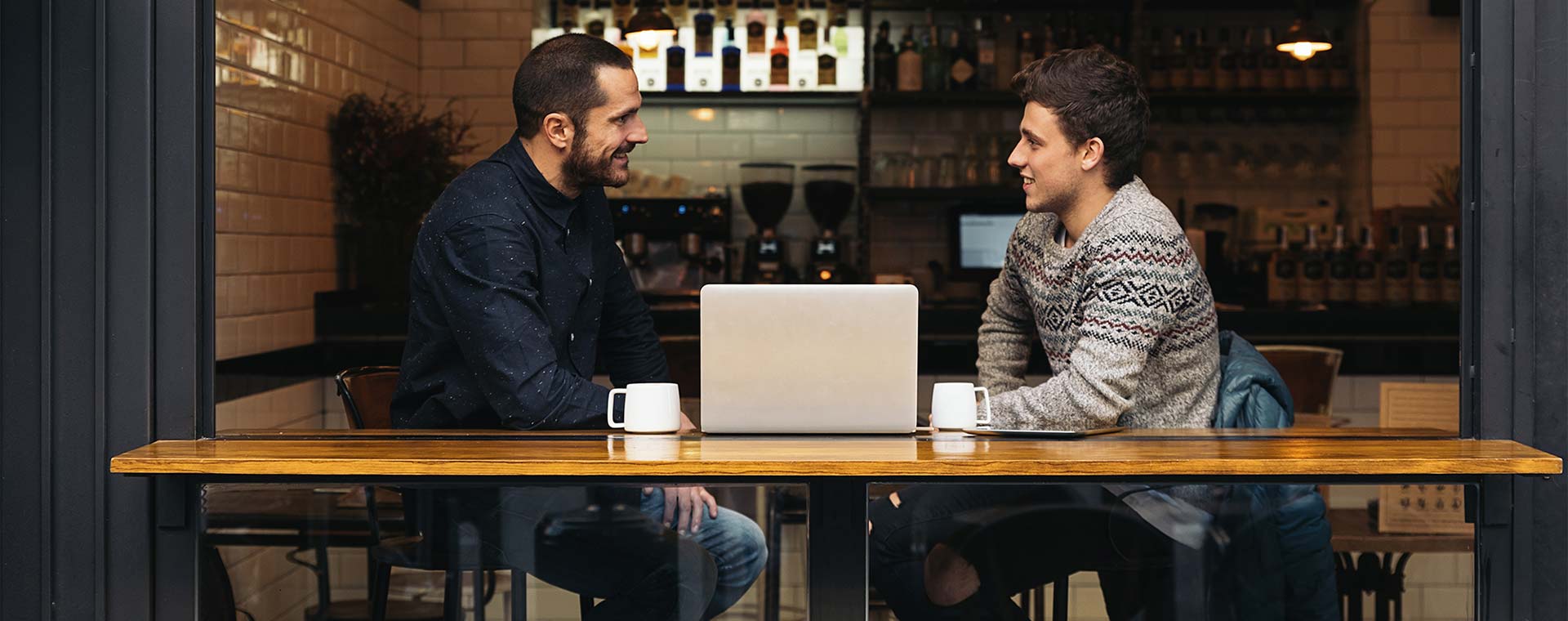 Two men are in a coffee shop sitting at a table, which looks out onto a street, talking to each other over a laptop. They each have a coffee mug, one is wearing a navy blue top and the other has a grey knit sweater on.