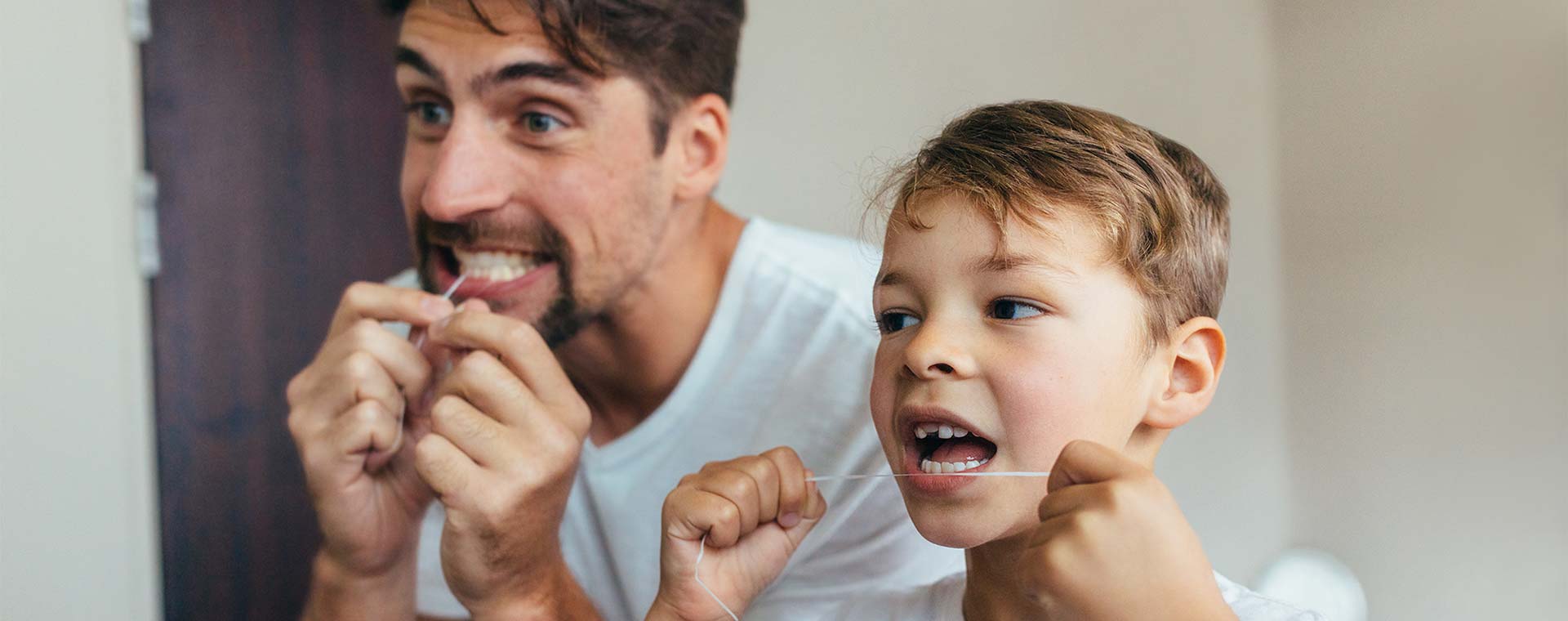 A father and son stand in front of a mirror and are flossing their teeth.