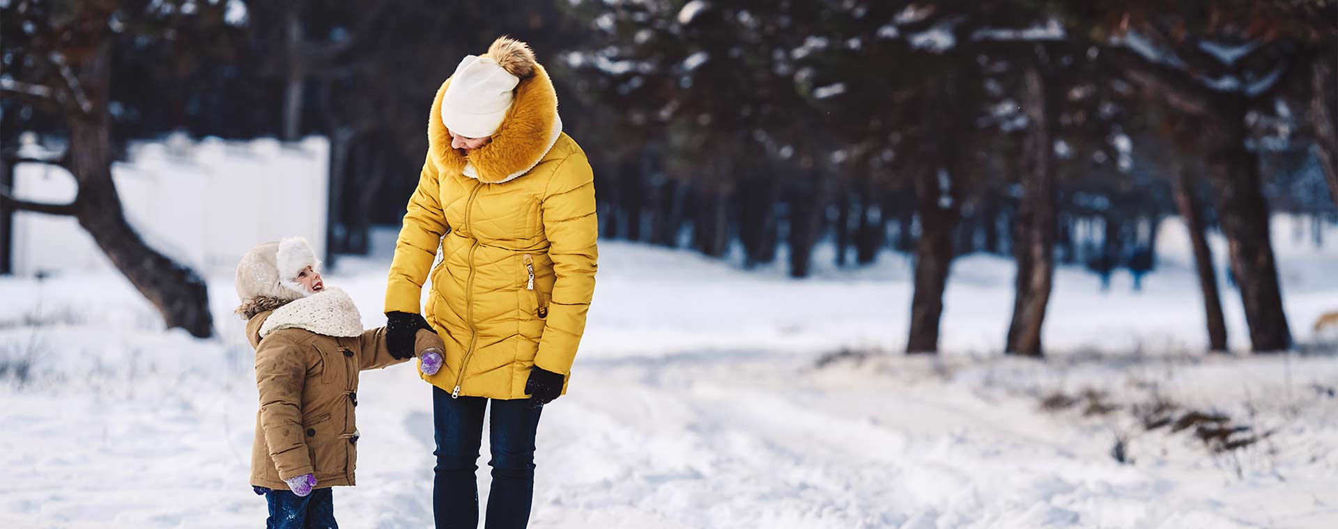 A mother in a yellow, puffy winter coat and her daughter in a light brown, puffy winter coat are walking in a forest. There is snow on the ground and in the trees.