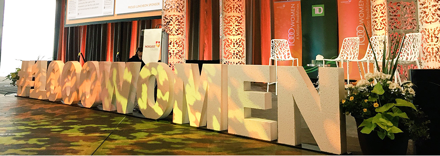 A side-on shot of a stage set to host a panel. There are black and red curtains handing behind the stage, with decorative screen just in front of the curtains. There are four white chairs, that have a back to match the screens, on the stage. In front of the stage in large, block Styrofoam letters it says Hashtag One Thousand Women.