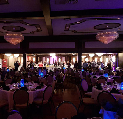 A view of a conference room with round tables set with white table clothes and dinner places. There are blue lights on each table. In the background is a group of people.