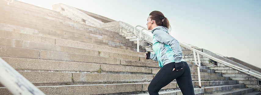 An attractive brunette female is running up concrete steps in early morning sunlight. She is wearing leggings, a jogging jacket and weighted gloves.