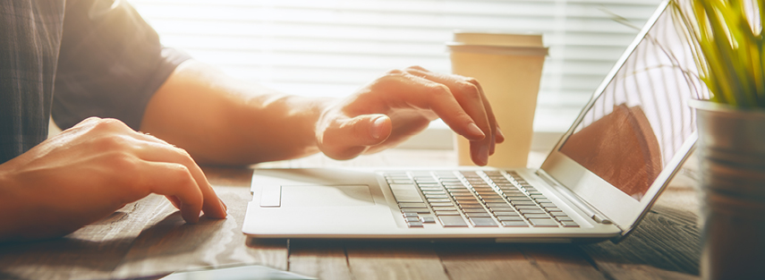 A side on, close up for someone sitting at a sleek laptop about to type on the keyboard. The are a disposable coffee cup beside the laptop and morning light is filtering through the window behind.