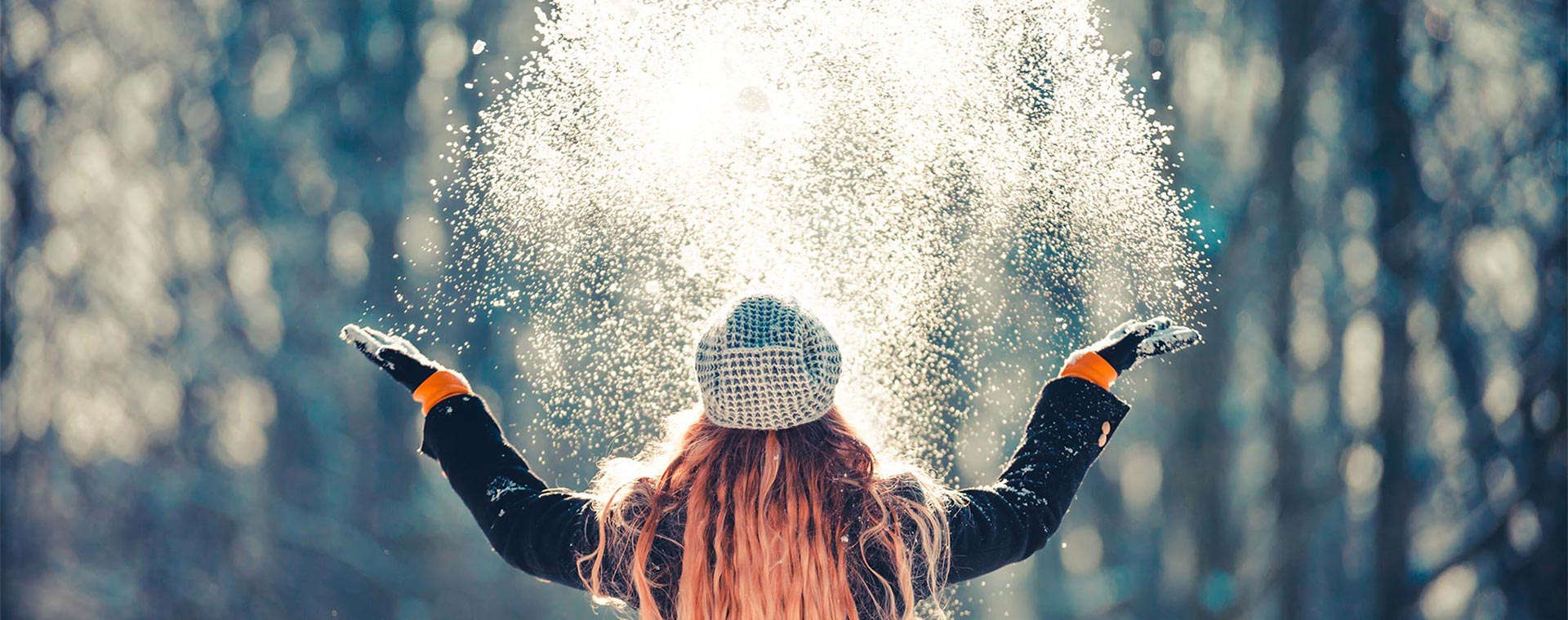 View from behind of a young woman with long, wavy, coral colored hair throwing snow up into the air with the sunlight shining through the snow, in front of trees
