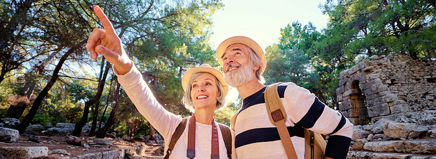 A couple in their early sixties are standing in front of ruins. There are many trees surrounding the ruins and the sun is just behind the couple. They are both wearing hats and backpacks, the woman has a camera around her neck.