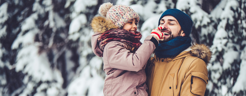 A father and daughter in warm winter coats, with fur hoods, are in a snow covered forest. The father is holding the daughter in his arms, while she boops him on the nose. They are both laughing.