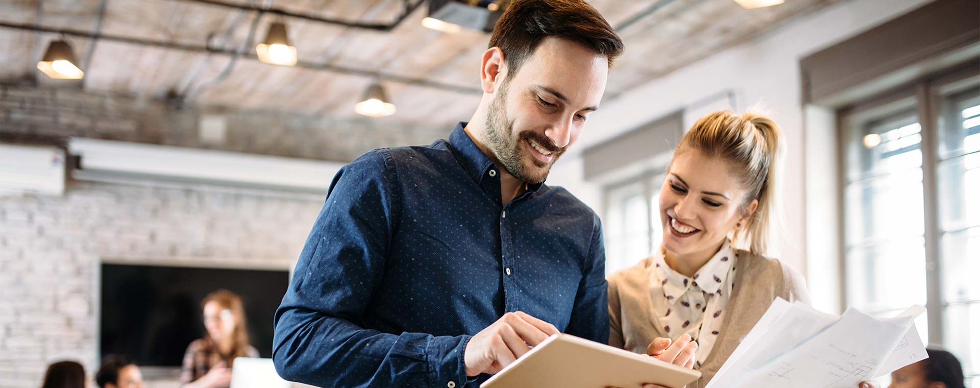 An attractive couple standing in a loft style office are smiling as they look down at a tablet the man is holding. He has a groomed stubble and well styled hair and is wearing a navy blue top. Her hair is pulled back into a pony tail and she is wearing a tan colored cardigan over a white collared shirt.