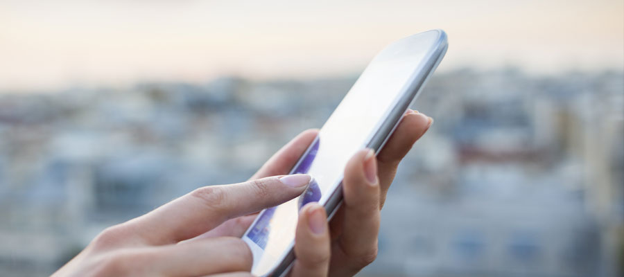 Side on close up of someone holding their phone at a slight angle, the reflection of light on the screen makes the screen barely visible. The background is a blurred out cityscape