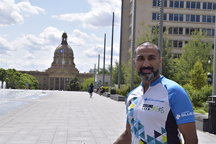 Sandeep in a Team Alberta Blue Cross cycling shirt standing near a fountain, with the Alberta Legislative Building in the background.
