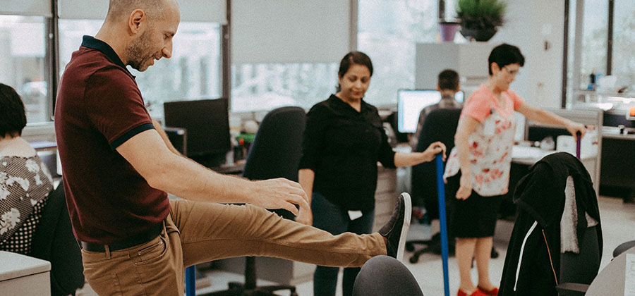 A multiracial group of business people in an open concept office, using items commonly found in the workplace to assist in stretching. In the background are two people sitting at their computers.