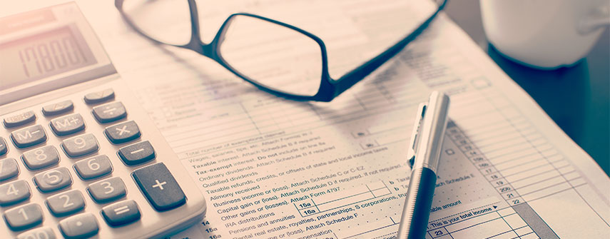 A close up of a tax form with a pen, a simple calculator and a pair of black rimmed glasses on top of the form.