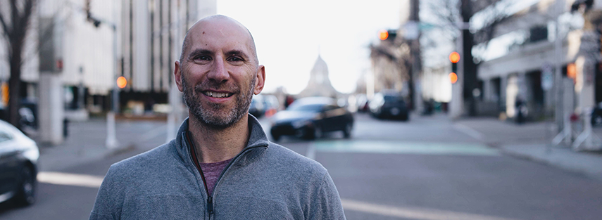 A head-shot of Terry Skidnuk standing in the middle of the road, with the street behind blurred. At the end of the street is the top of the Alberta Legislative Building.