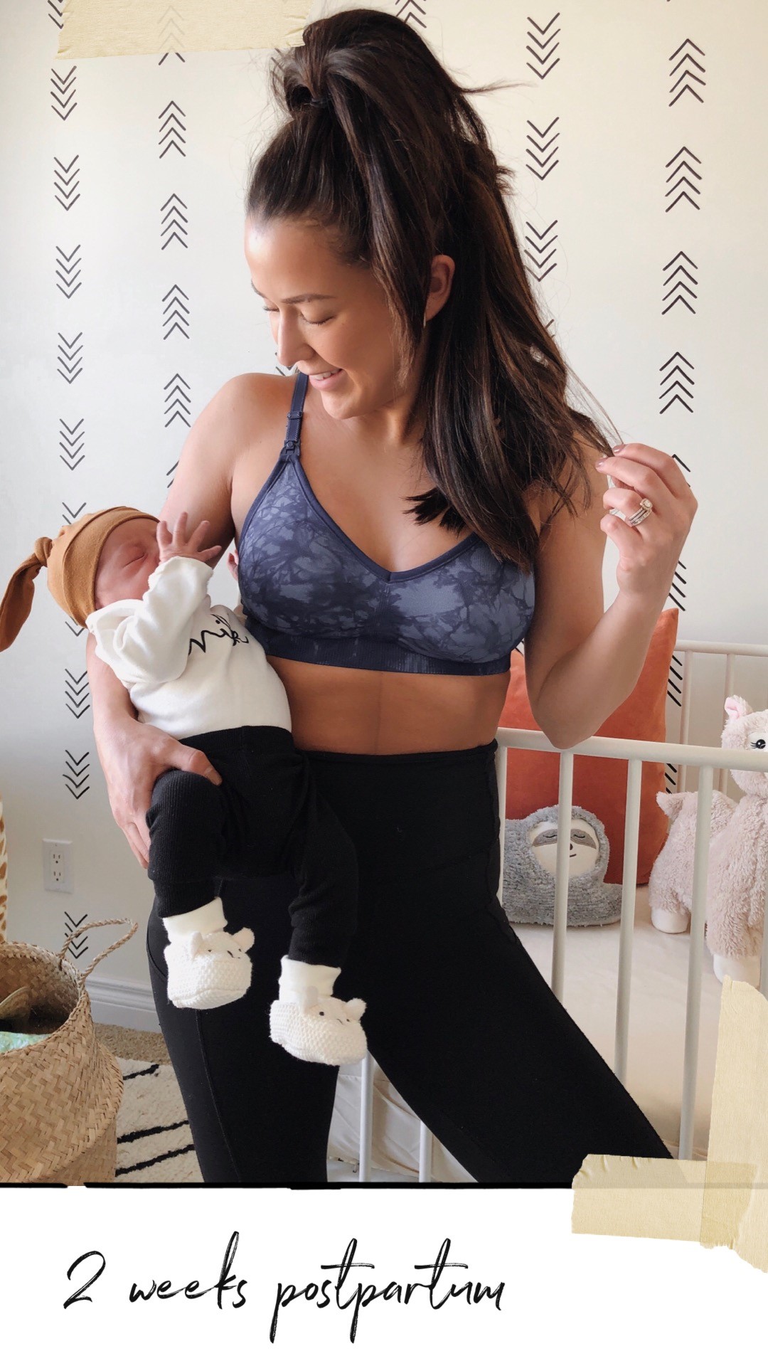 Lindsay Smith wears leggings and a sports bra while she holds her infant, looking down and smiling. 