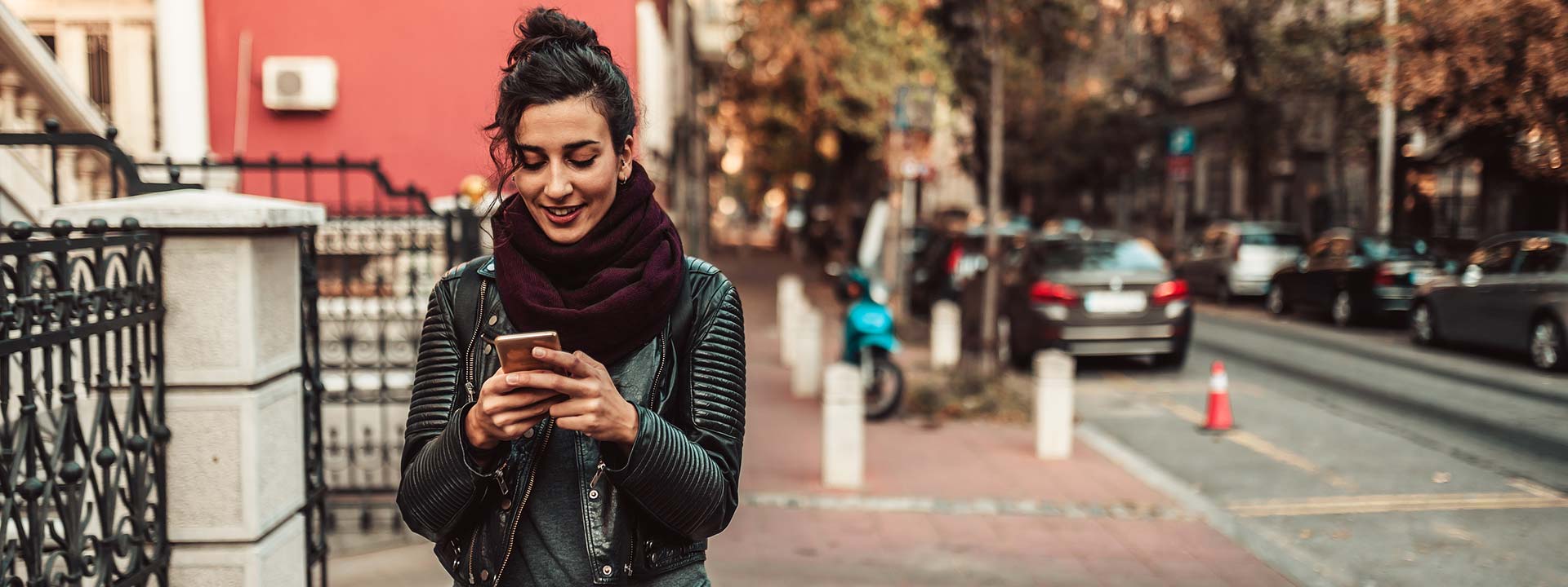 A young woman is walking down a street, looking down at her phone and smiling.