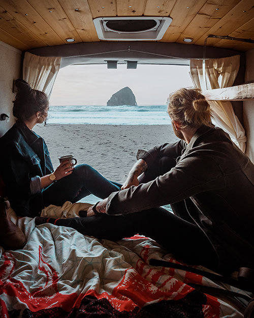 Jay R. Mcdonald and a women sit in the back of a van, drinking coffee and looking out over a beach that leads to the ocean.