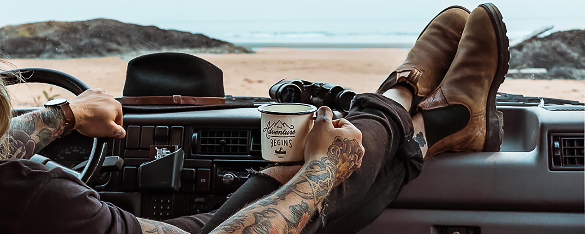 Jay R. Mcdonald sits in the driver's seat of a car with his feet up on the dash and a mug of coffee in his hand. in the distance there is a beach leading to the ocean.