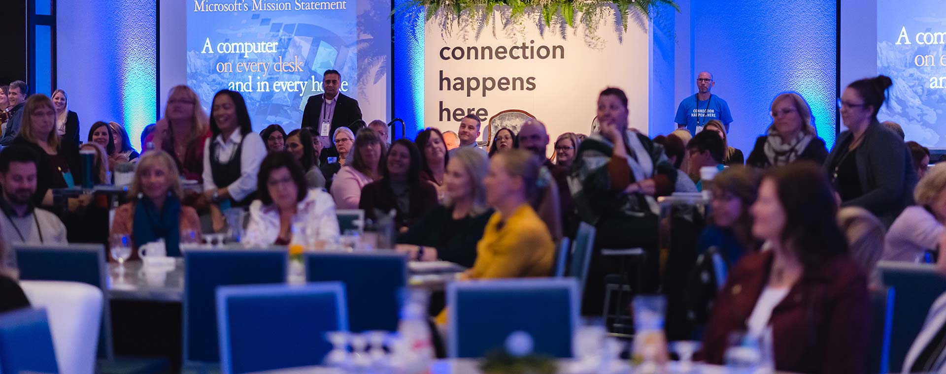 A large room filled with people who are listening to a guest speaker. The room has ambient blue lighting and greenery over a sign that reads connection happens here.