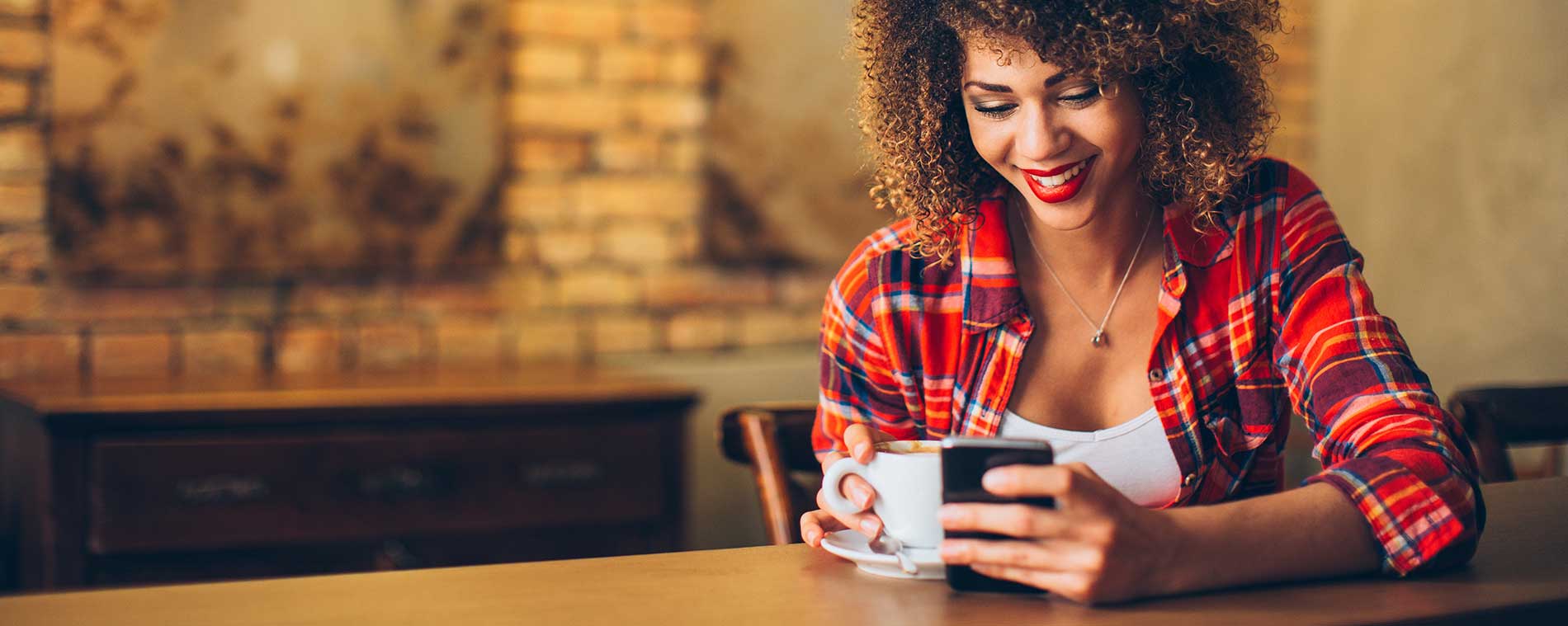 A young woman is sitting in a coffee shop, cup in hand, smiling and looking down at her smartphone.