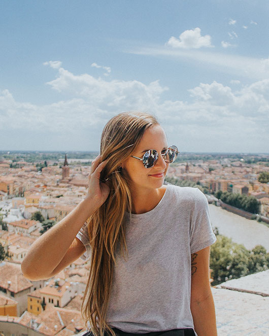 ZandaLee sitting on a wall in an ancient city, she is wearing reflective sunglasses and a light grey tshirt.