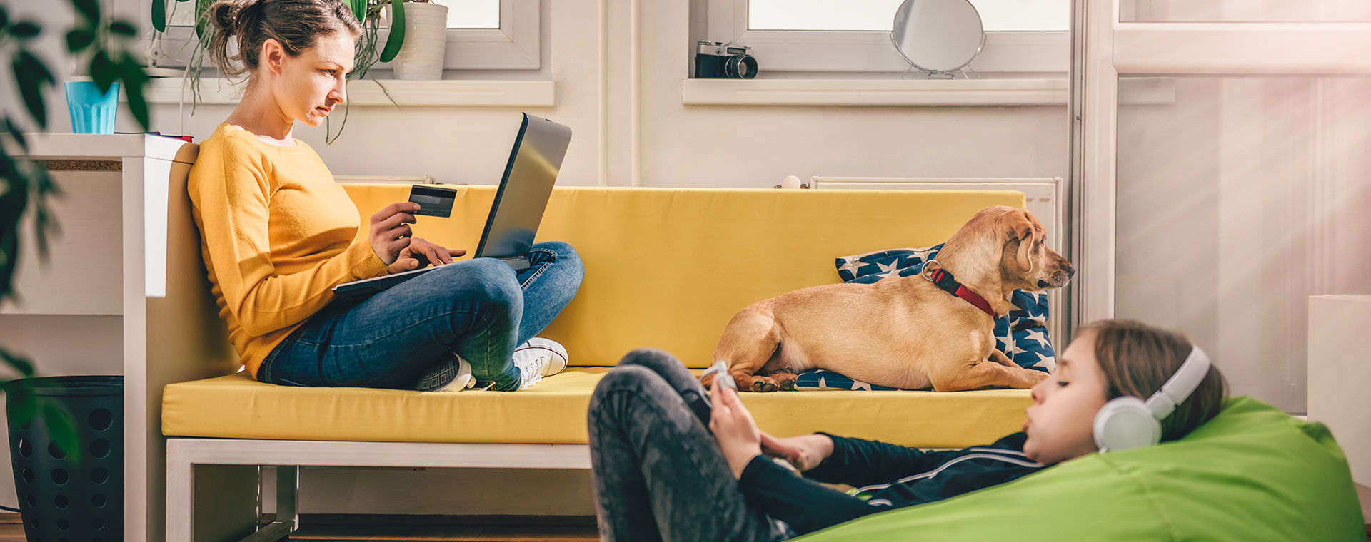 A young mother is shopping on a laptop, she has her credit card in one hand. On the couch beside her is a golden lab puppy. In the foreground is a young a child, looking at a smartphone where headphones.