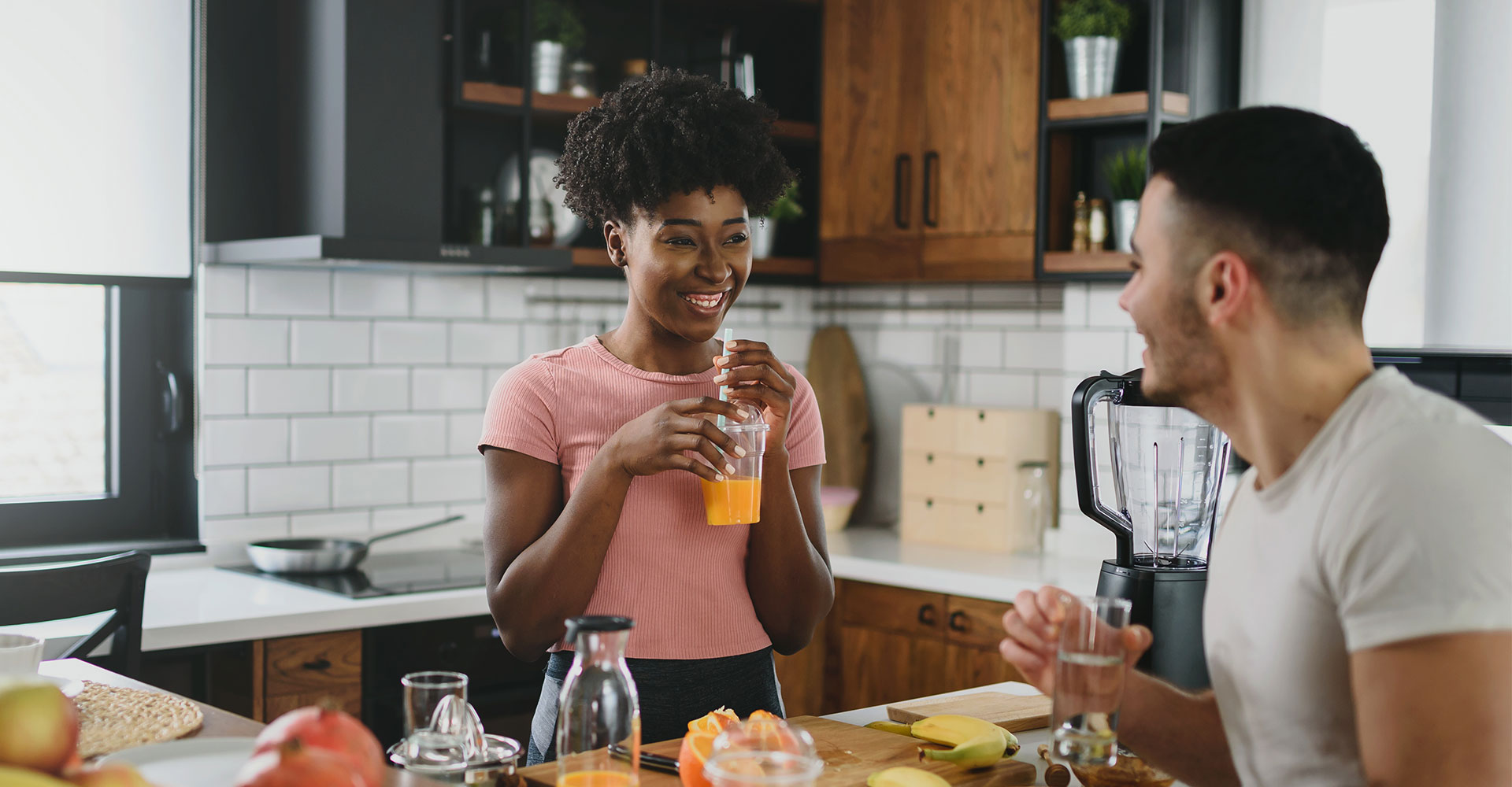 A young, happy, multiracial couple are have eating breakfast in a modern-farmhouse style kitchen. They have lots of fruit on the counter and are drinking fresh orange juice.
