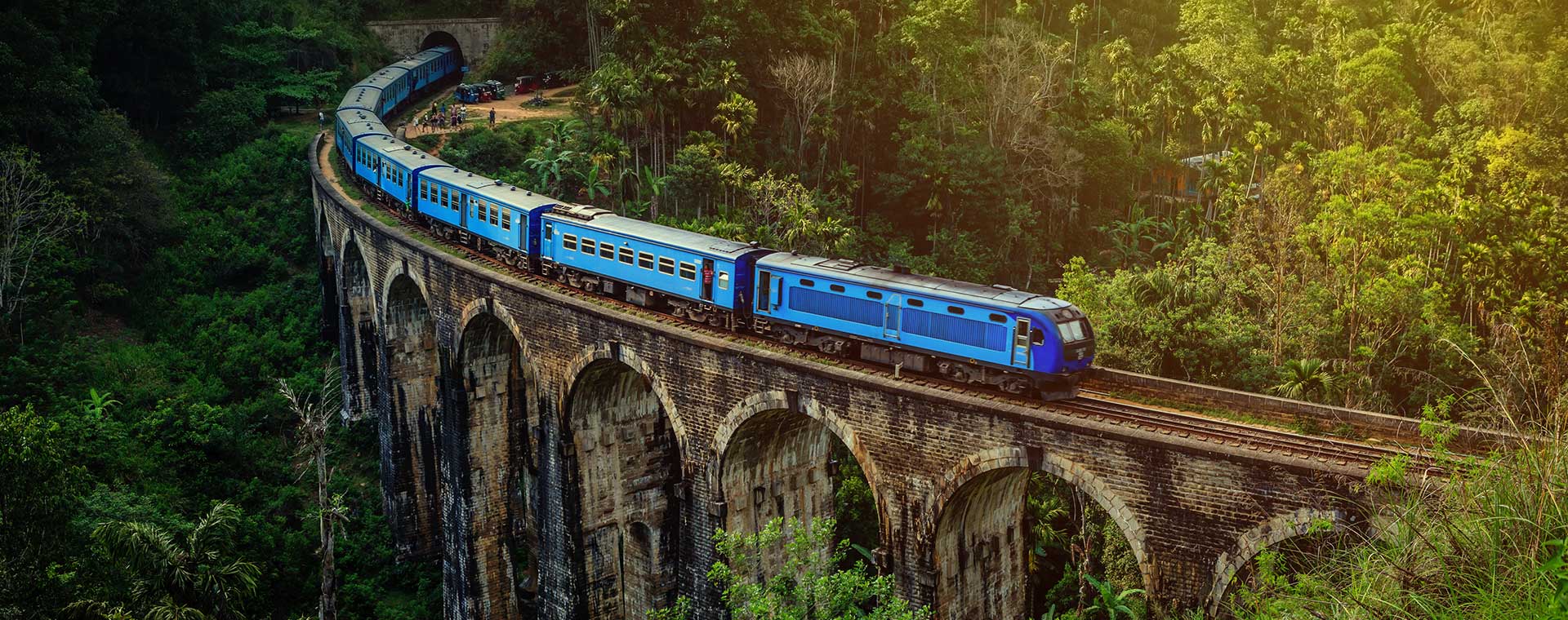 A blue train is crossing the Nine Arches Bridge, in Sri Lanka. The bridge is surrounded by lush green rainforest. There is soft, yellow sun light filtering through from the top right corner of the image.