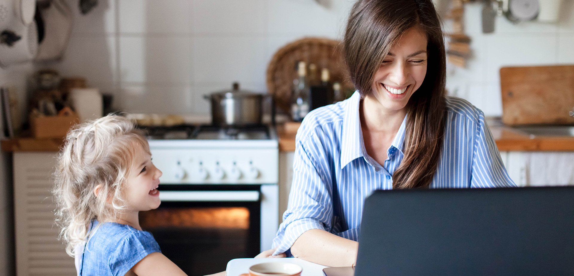Working mom works from home office. Happy mother and daughter smiling. Successful woman and cute child using laptop. Freelancer workplace in cozy kitchen. Female business. Lifestyle authentic moment.