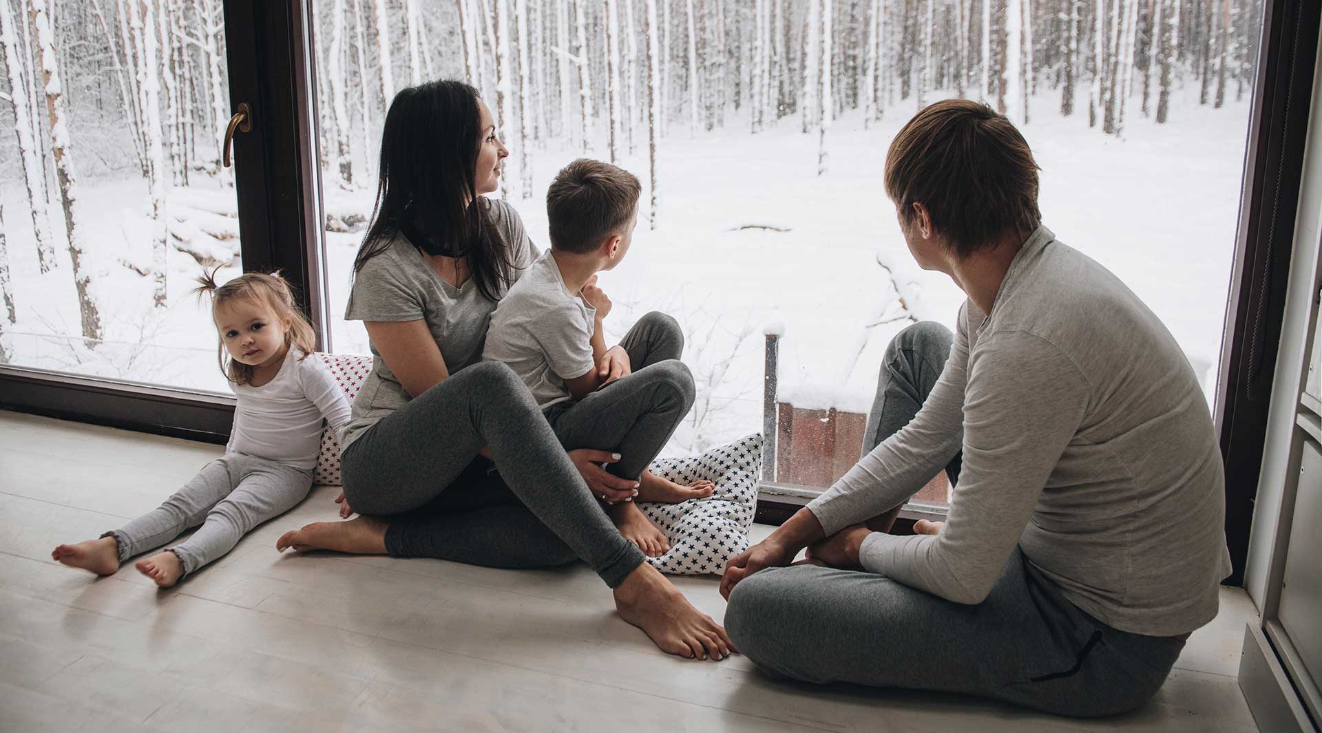 A family of four are sitting on their floor, looking out a large glass window at a wintry backyard.