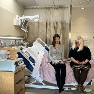 Two women reading book while sitting on hospital bed.