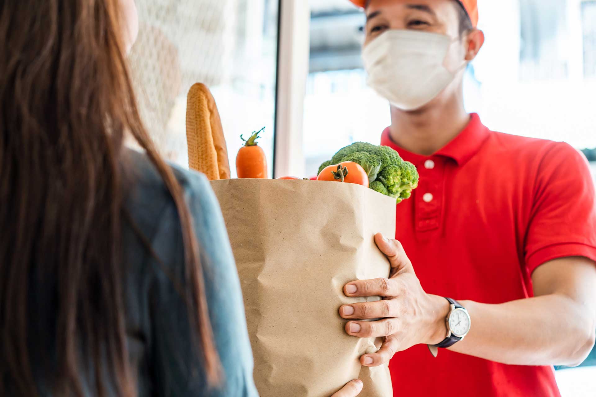 Delivery man wearing face mask in red uniform handling bag of food, fruits, vegetables to female costumer in front of the house.
