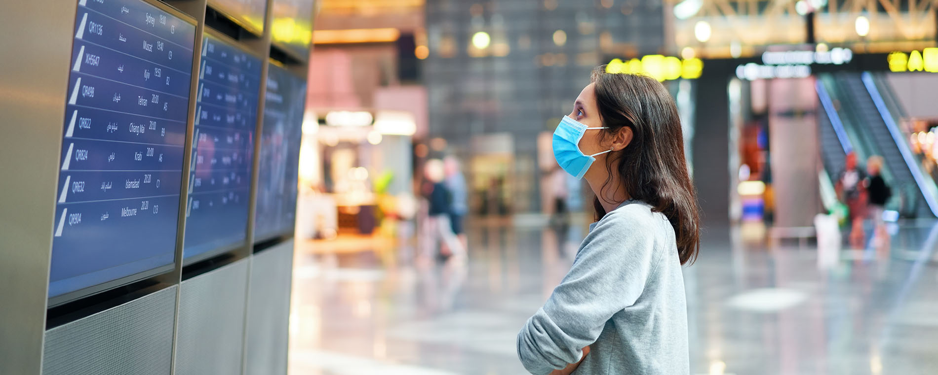 Woman in virus protection face mask looking at information board checking her flight in international airport. Departure board, flight status