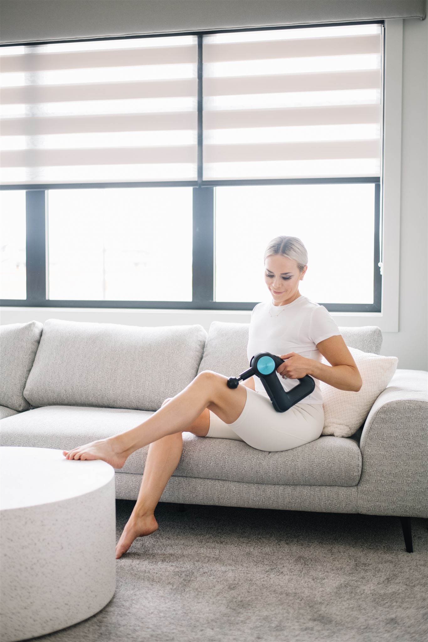 Richelle sitting on her couch using a Theragun on her leg.