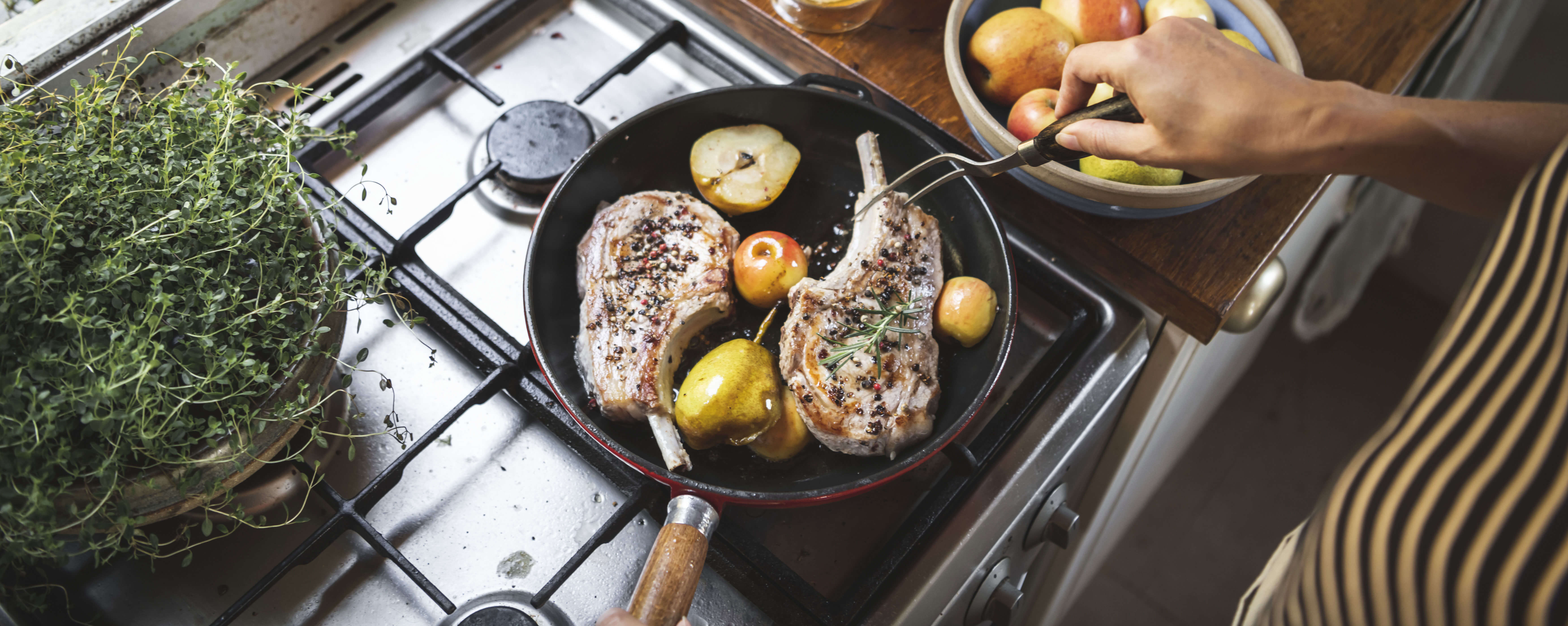 A person is cooking pork chops on a propane stovetop. In the frying pan are fresh herbs, apple and pear halves.