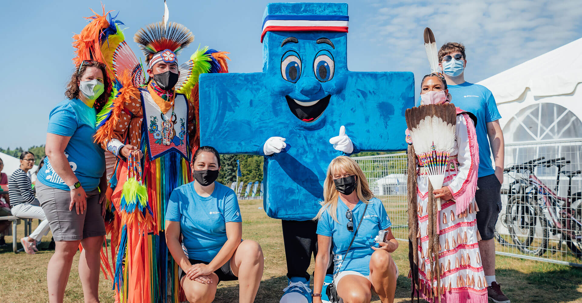 Alberta Blue Cross mascot, Big Blue is standing with two First Nations dancers dressed in vibrant colours. There are members of the Alberta Blue Cross Street Team with them.