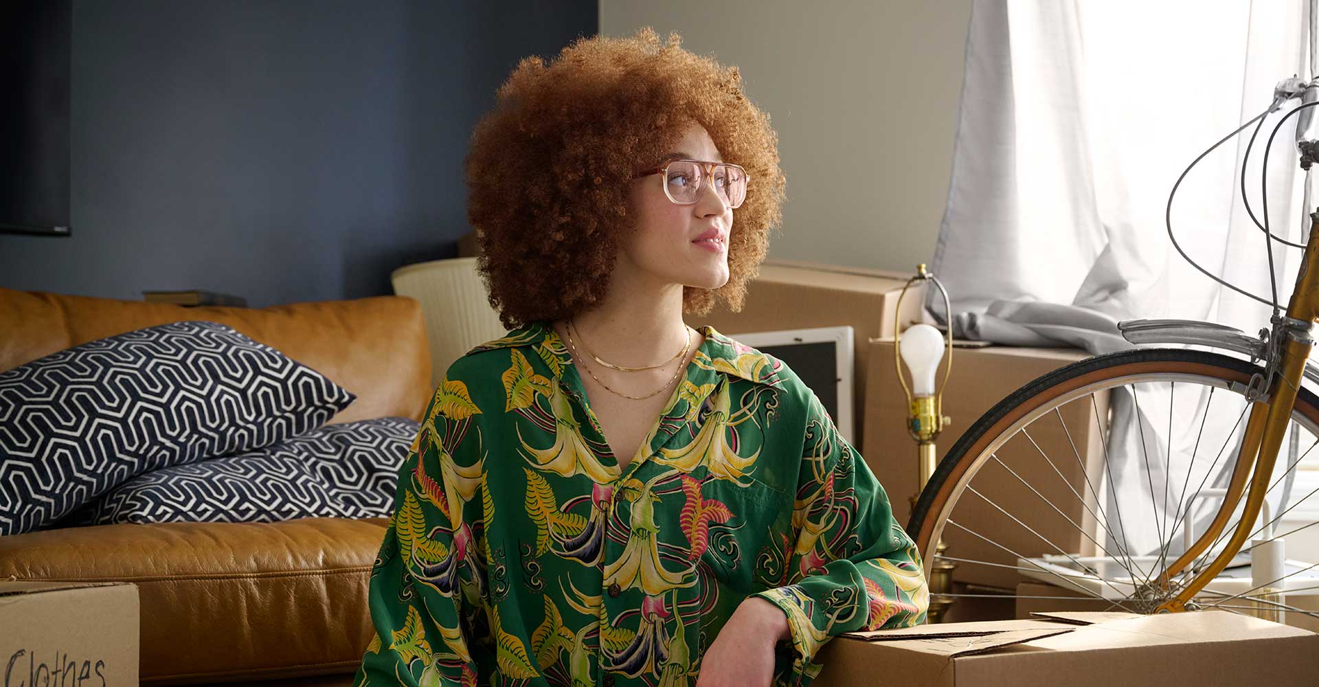 Young woman wearing 70's style glasses and green floral blouse sitting on an apartment floor and looking out a window while surrounded by moving boxes and a bicycle.