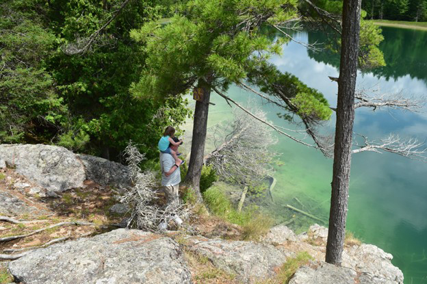 A man with a kid sitting on his shoulder standing on rocks overlooking a lake.