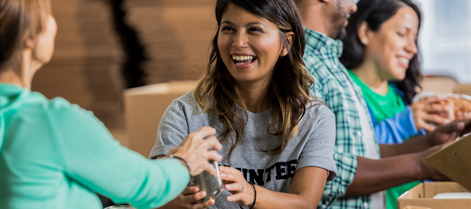 A woman smiles while working with other volunteers at a local food bank. They are packing cardboard boxes with canned goods to be sent to people in need.