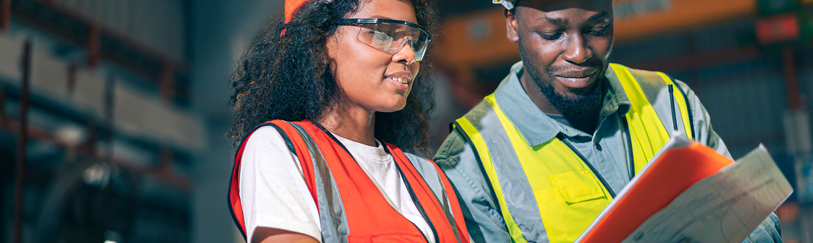 Two professional engineer/worker/technician use clipboard discuss work, walk in steel metal manufacture factory plant industry. Black African American man and woman wear hard hat check quality machine