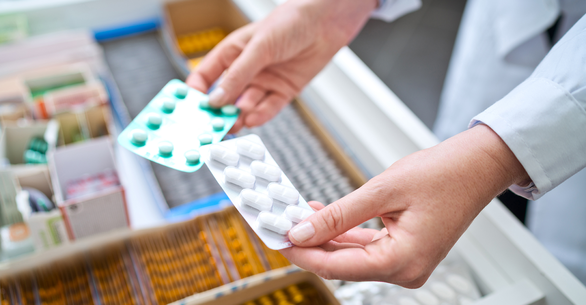 Pharmacist holding blister packs of white and green pills, with a drawer full of medications in the background. illustrating drug pipeline.