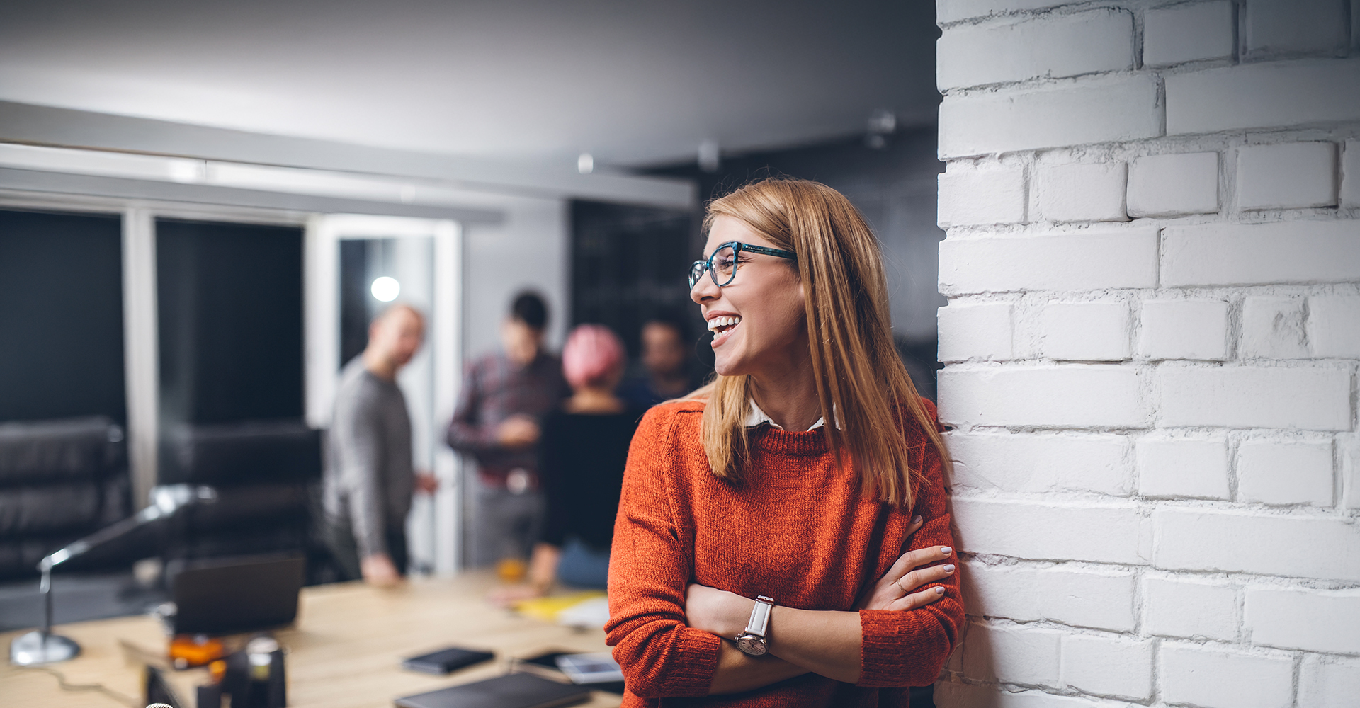 Person in an orange sweater leaning against a white brick wall in a modern office setting with colleagues in the background.