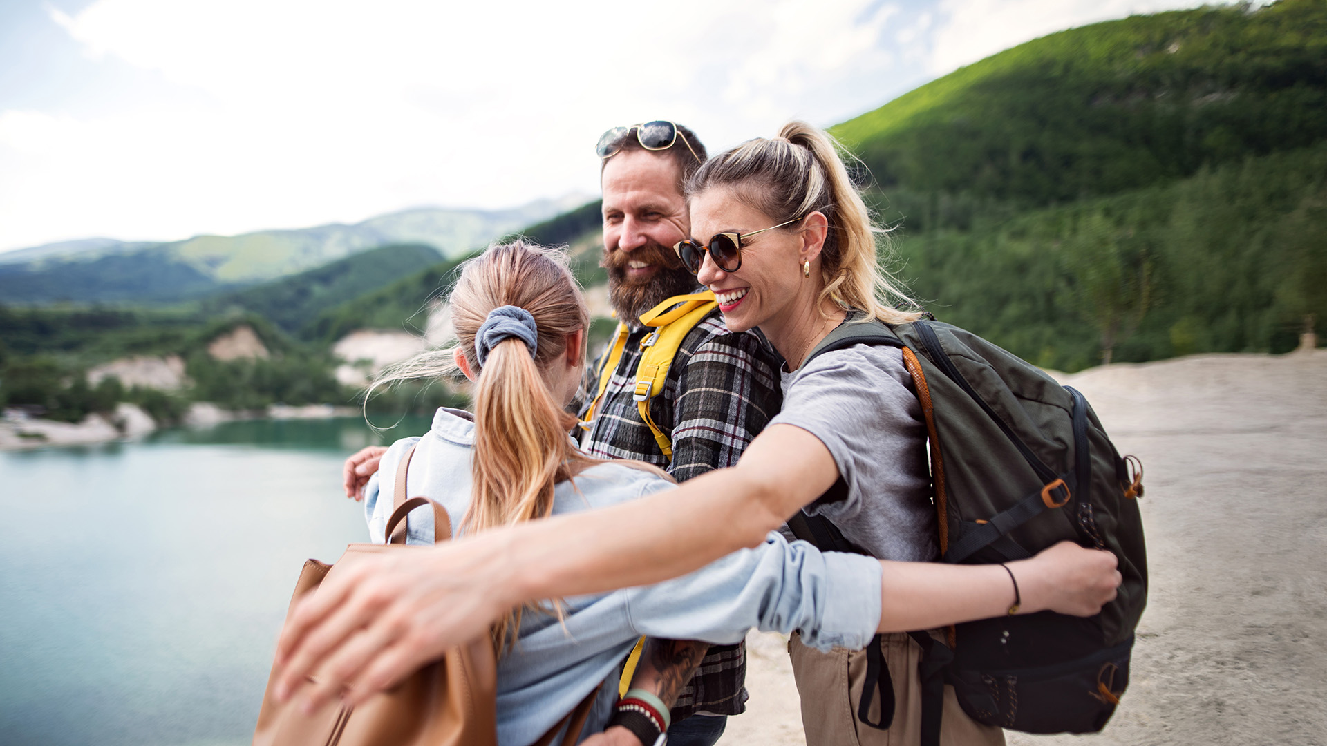 A smiling family of three share a group hug while hiking near a scenic mountain lake. They are wearing backpacks and casual outdoor clothing, with forested hills and calm water in the background.