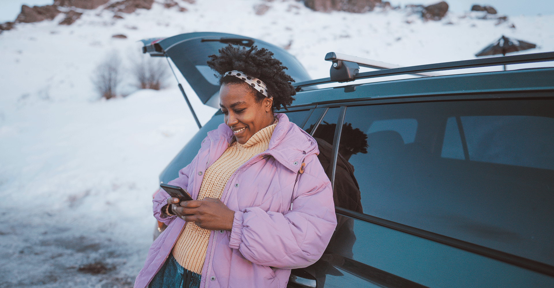 A young traveller stands beside an open car trunk in a snowy mountain setting, checking their phone as part of their travel planning.